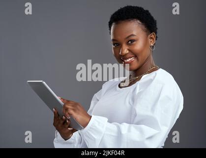 Mit einem einfachen Klick können Sie Ihre Träume wahr werden lassen. Eine junge Frau mit einem digitalen Tablet, während sie vor einem grauen Hintergrund steht. Stockfoto