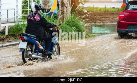 Frau fahren Motorrad durch überflutete Straße. Motorradfahren auf überfluteter Straße während der Überschwemmung durch sintflutartige Regenfälle. Überflutete Straße mit großen Stockfoto