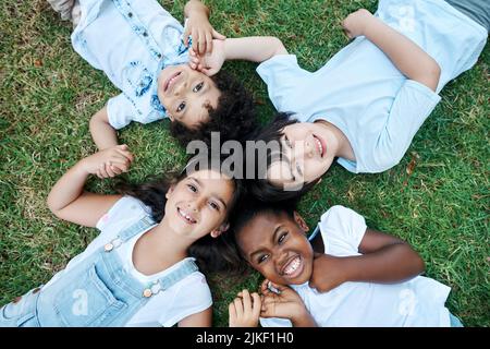 Es ist immer ein toller Tag, wenn ich meine Freunde um mich habe. Vier entzückende Kinder liegen zusammen auf dem Rasen. Stockfoto