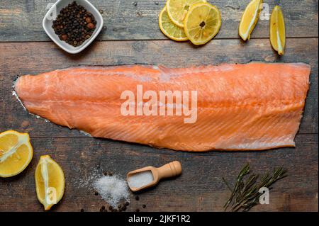 Rohe Lachsfilets mit Zitrone, Salz, Pfeffer. Gekühltes Lachsfilet auf der Haut als knochenlose Hälfte des Fisches, liegt die Haut mit der Seite nach unten auf rustikalem Holzhintergrund Stockfoto