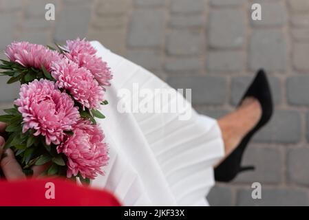 Bouquet von dunkelrosa Astern in den Händen einer sitzenden Frau in einem weißen Faltenrock und schwarzen Stilettos, Draufsicht. Konzept der Entspannung und Reflexion. Stockfoto