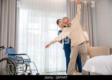 Der alte ältere Mann trainiert gerne bei einem Physiotherapeuten für ausgestreckte Arme zu Hause, der asiatische Physiotherapeut-Patient stillt ältere Menschen beim Trainieren Stockfoto