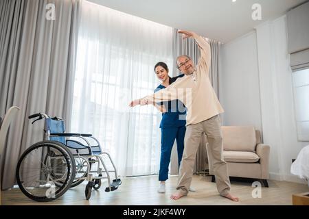 Der alte ältere Mann trainiert gerne bei einem Physiotherapeuten für ausgestreckte Arme zu Hause, der asiatische Physiotherapeut-Patient stillt ältere Menschen beim Trainieren Stockfoto