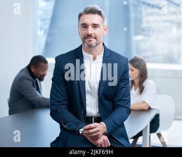 HES ist als echter Marktführer in der Branche bekannt. Porträt eines selbstbewussten, reifen Geschäftsmannes, der in einem Büro mit seinen Kollegen im Hintergrund steht. Stockfoto