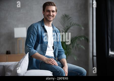 Porträt eines attraktiven charismatischen kaukasischen Mannes in einem Jeanshemd, der auf einem Sofa im Wohnzimmer sitzt und mit einem freundlichen Lächeln auf die Kamera blickt Stockfoto