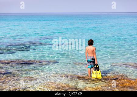 Junge, der an einem Ufer mit Schnorchelflossen in den Händen an einem wunderschönen Strand von Veli ZAL, Dugi otok, Kroatien, steht Stockfoto