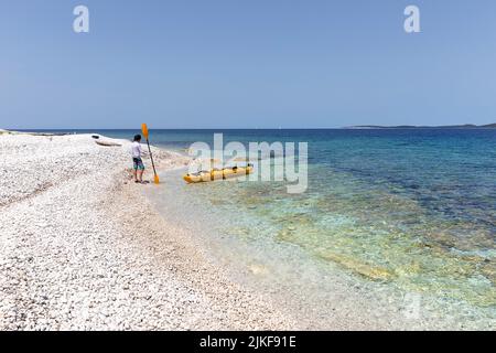 Junge steht auf einem Kiesstrand in der Nähe eines Seekajaks auf einer unbewohnten Insel Mezanj in der Nähe des Strandes Veli ZAL, Insel Dugi otok, Kroatien Stockfoto