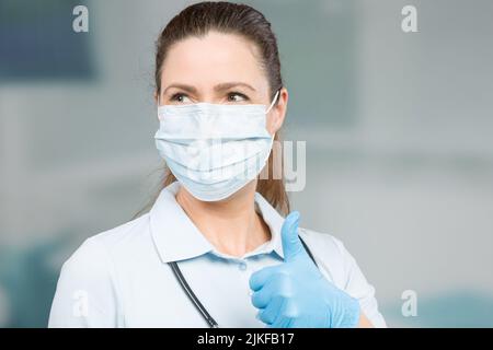 Ärztin mit medizinischer Gesichtsmaske und Stethoskop zeigt Daumen nach oben Stockfoto