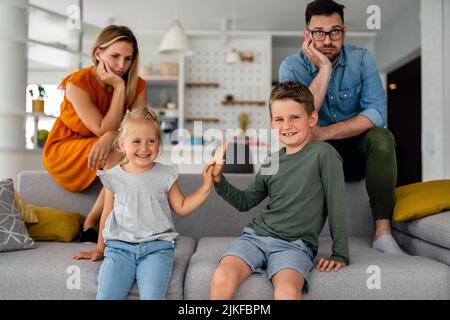 Müde Eltern, die auf der Couch sitzen, fühlen sich erschöpft genervt, während glückliche Kinder zusammen spielen. Stockfoto