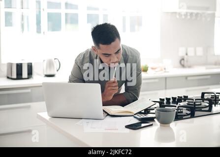 Ein junger Mann, der einen Laptop benutzt, während er Notizen in der Küche zu Hause durchläuft. Stockfoto