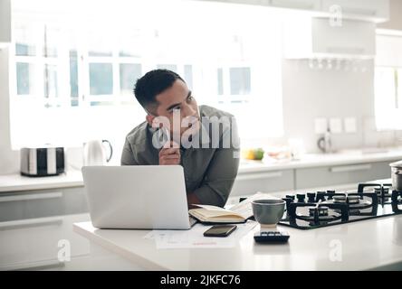 Ein junger Mann mit einem Laptop, während er Notizen in der Küche zu Hause durchläuft. Stockfoto