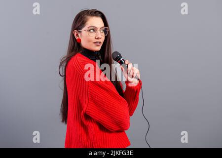 Junge, emotionale Brünette mit einem Mikrofon, das in einem roten Pullover gekleidet ist, singt Karaoke oder sagt eine Rede, isoliert auf Grau Stockfoto