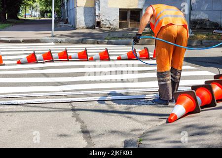 Ein Straßenarbeiter in orangefarbenen Overalls zeichnet weiße Straßenmarkierungen eines Fußgängerübergangs auf einem eingezäunten Abschnitt der Fahrbahn. Speicherplatz kopieren. Stockfoto