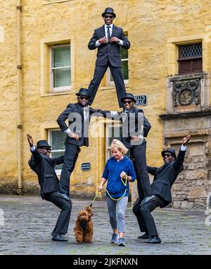 Edinburgh, Schottland, Großbritannien, 2.. August 2022. Edinburgh Festival Fringe: Die Black Blues Brothers. Diese Gruppe afrikanischer Akrobaten, die zuletzt 2019 im Fringe auftraten, zeigt ihre Akrobatik im Dean Village während des Edinburgh Festival Fringe. Im Bild:mDie ortsansässige Susie Macleod geht mit ihrem Labradoodle-Hund unter die Pyramide Stockfoto