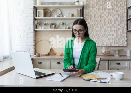 Seriöse und fokussierte Geschäftsfrau freiberuflich auf Papier arbeiten zu Hause in der Küche, brünett in der Brille und grün in der Küche, zahlen und Rechnungen zu berechnen Stockfoto