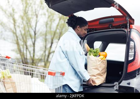 Seitenansicht Porträt einer jungen schwarzen Frau, die auf dem Supermarkt-Parkplatz eine Einkaufstasche in den Kofferraum legt, Kopierplatz Stockfoto