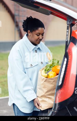 Vertikales Porträt einer jungen schwarzen Frau, die auf dem Supermarkt-Parkplatz eine Einkaufstasche in den Kofferraum legt Stockfoto