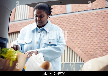 Waist-up-Porträt einer echten jungen Frau, die nach dem Einkaufen im Supermarkt Einkaufstasche in den Kofferraum legt Stockfoto