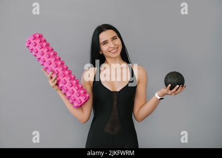 Eine attraktive schlanke Frau in sportlicher schwarzer Kleidung mit einem Massageball und einer Rolle in den Händen lächelt auf einem grauen Hintergrund. Fitnesstrainer Stockfoto