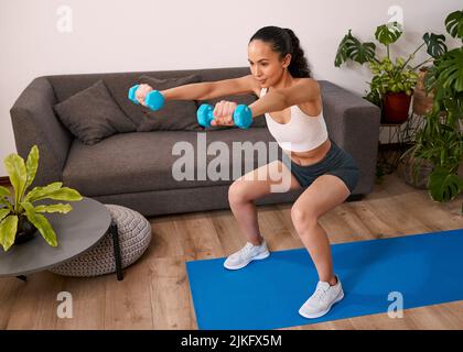Eine junge Frau hockt und hebt Gewichte zu Hause, um fit zu werden Stockfoto