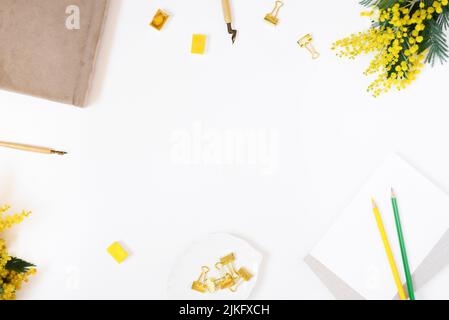 Ein moderner Schreibtisch für das Home Office eines Bloggers oder Studenten mit einem Notizbuch aus Papier, einem Stift für Kaligraphie, Buntstiften und Mimosenblumen auf einem weißen Backgroun Stockfoto