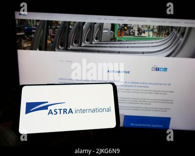 Person, die Mobiltelefon mit Logo der indonesischen Firma PT Astra International Tbk auf dem Bildschirm vor der Webseite hält. Konzentrieren Sie sich auf die Telefonanzeige. Stockfoto