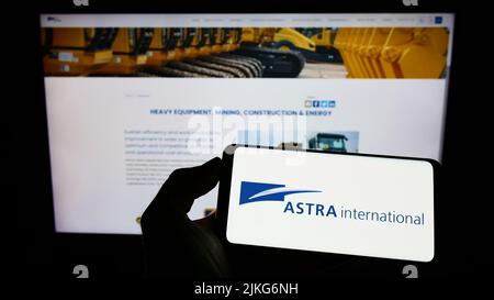 Person, die Mobiltelefon mit Logo der indonesischen Firma PT Astra International Tbk auf dem Bildschirm vor der Webseite hält. Konzentrieren Sie sich auf die Telefonanzeige. Stockfoto