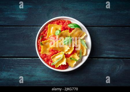 Ravioli mit Tomatensauce und frischen Basilikumblättern auf einem Teller, über dem Kopf flach auf dunklem Holzhintergrund aufgelegt Stockfoto