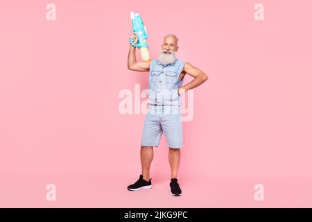 In voller Länge Foto von attraktiven starken Mann Rentner gekleidet Denim Weste steigenden Wasserpistole isoliert rosa Farbe Hintergrund Stockfoto