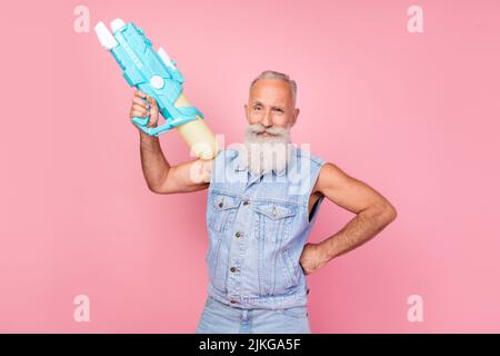 Foto von coolen attraktiven Mann Rentner gekleidet Denim Weste halten Wasserpistole isoliert rosa Farbe Hintergrund Stockfoto