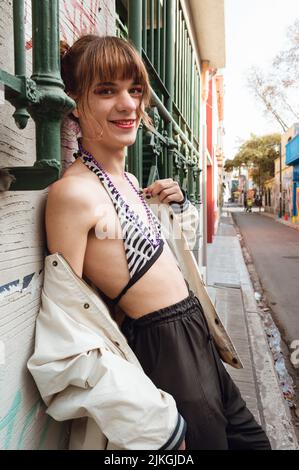 Die transsexuelle junge lateinische kaukasische Frau mit rotem Make-up und spärlich bekleideter Verkleidung ist lächelnd an der Wand gelehnt und blickt in p auf die Kamera Stockfoto