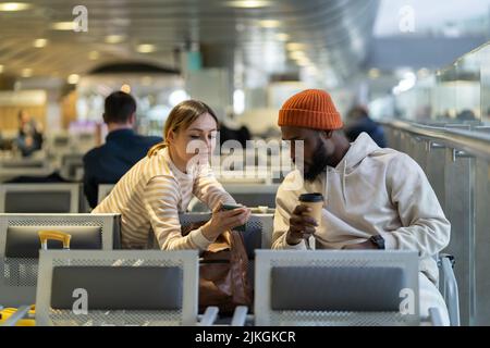 Schwarzer Hipster, männlich und junge kaukasische Frau, die beim Warten auf den Flug am Flughafen auf das Telefon schaut. Stockfoto