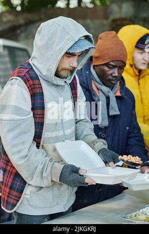 Gruppe von Obdachlosen in warmer Kleidung, die mit Plastiktellern standen und auf die Abgabe von Nahrungsmitteln durch Freiwillige warteten Stockfoto