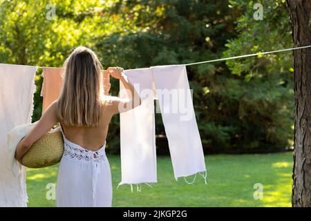 Rückansicht einer nicht erkennbaren jungen blonden Frau im Boho-Outfit, die im Sommer die Wäsche im Garten aufgehängt hat, mit Platz für Kopien Stockfoto