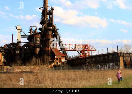 Eine Frau mit einem Koffer, die in der Nähe einer rostigen alten Stahlfabrik auf einem Feld läuft Stockfoto
