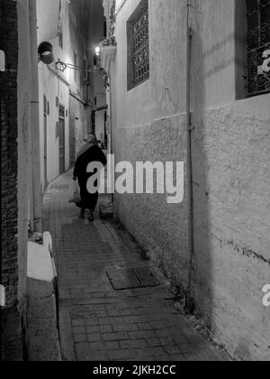 Eine vertikale Graustufenaufnahme einer Frau, die durch eine Gasse in Tanger, Marokko, geht Stockfoto