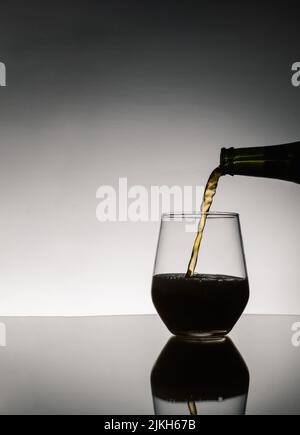 Das Bier wurde aus der Flasche, die auf dem Tisch auf dunklem Grund gespiegelt war, in ein Glas gegossen Stockfoto