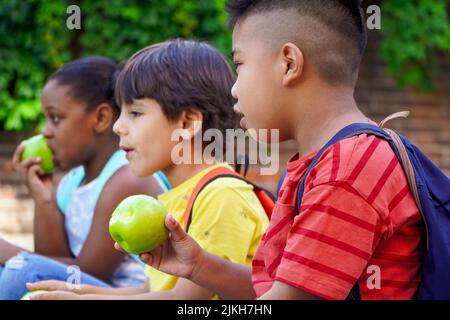 Multiethnische Kinder mit Rucksäcken sitzen auf der Straße am Schuleingang und essen Äpfel. Zurück zum Schulkonzept. Multiethnische Kindergruppe Stockfoto