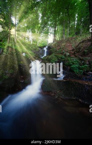 Eine vertikale Aufnahme von Wasserfällen, die die Steine hinunterstürzen, mit Sonnenlicht, das durch die Bäume im Hintergrund scheint Stockfoto