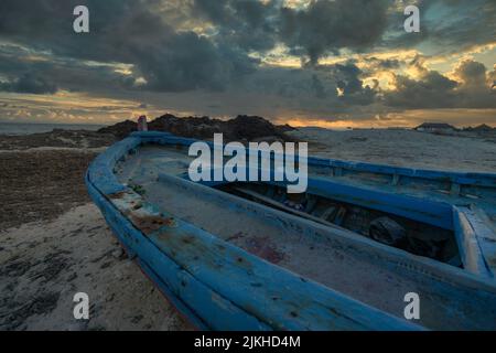 Eine schöne Aufnahme eines blau rostigen Bootes auf einem Sand mit Sonnenuntergang im Hintergrund Stockfoto