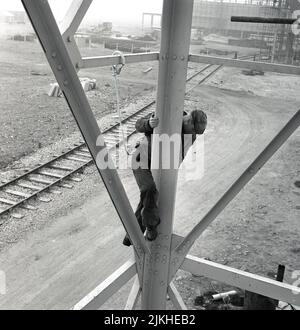 1950s, historisch, ein Arbeiter in einem großen Stahlwerk, hoch über dem Boden kletternd auf der Außenseite einer Stahlkonstruktion, mit einem Sicherheitsseil an ihm befestigt, Stoffkappe auf dem Kopf, Abbey Works, Port Talbot, Wales. VEREINIGTES KÖNIGREICH. Stockfoto