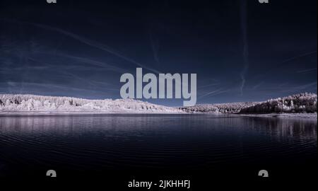 Ein Panoramablick auf den blauen See in der Nähe des schneebedeckten Waldes unter dem dunklen Himmel Stockfoto