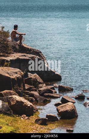 Ein junger Kaukasusmann, der auf dem Felsen sitzt und auf das Meer blickt Stockfoto