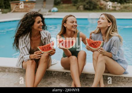 Drei niedliche junge Frauen sitzen am Pool und essen Wassermelone im Hinterhof des Hauses Stockfoto