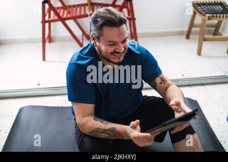 Ein lächelnder kaukasischer Mann mit einem Tablet, der zu Hause auf einer schwarzen sportlichen Yogamatte sitzt Stockfoto