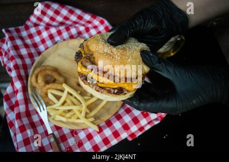 Eine Nahaufnahme der Hände, die einen Burger im Hintergrund von Zwiebelringen und pommes auf einem Holzteller halten. Stockfoto