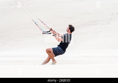 Junger Mann, der zwei Kontrollgriffe eines fliegenden Quad-Lines-Trainers übernimmt Parafoil / 4-Line-Stunt-Kite am Sandstrand bei starkem Wind im Sommer Stockfoto