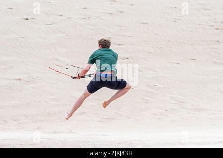 Junger Mann, der im Sommer bei starkem Wind an einem fliegenden Quad-Lines-Trainer Parafoil 4-Line-Control-Bar umwickelt / 4-Line-Stunt-Kite am Sandstrand Stockfoto