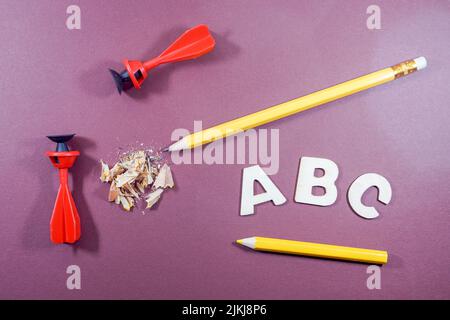 Eine Draufsicht auf ABC-Buchstaben in Weiß, Bleistift, Spitzerrückstand, gelber Farbstift und zwei Saugerabnäher auf violettem Hintergrund Stockfoto