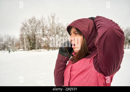 Eine Nahaufnahme einer überraschenden kaukasischen Frau, die mit Schnee und Bäumen um sich telefoniert